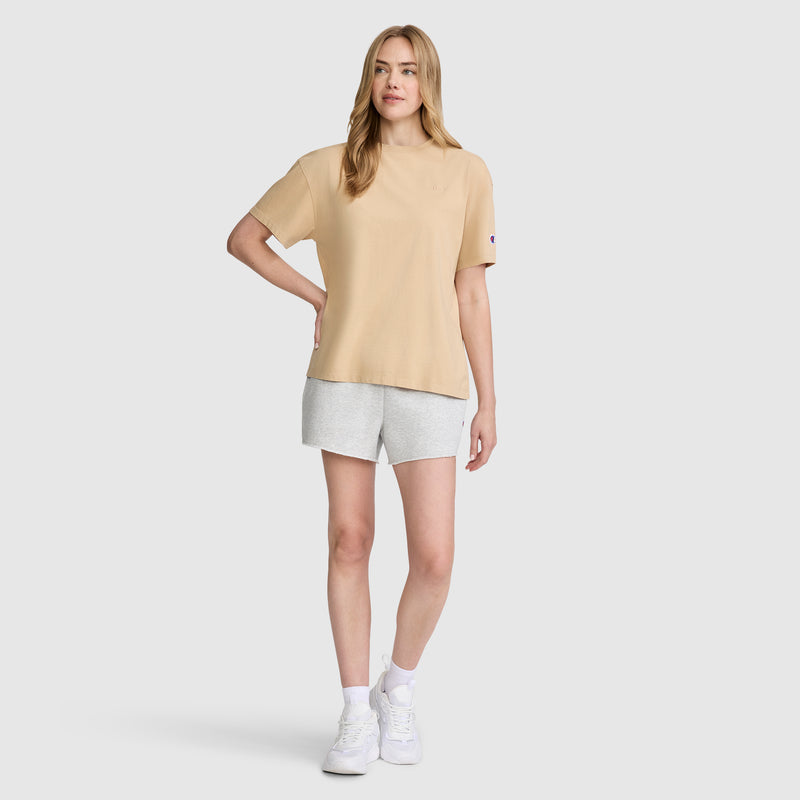 A woman stands against a plain background wearing the Champion Short Sleeve Loose Fit T-Shirt in beige cotton jersey, paired with light gray shorts and white sneakers. She poses with one hand on her hip and a relaxed expression.