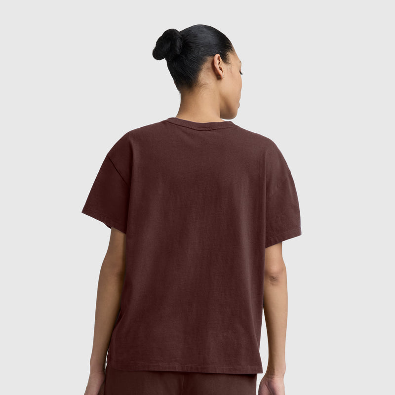A person with dark hair in a bun is seen from the back, wearing the Champion Short Sleeve Loose Fit T-Shirt in brown along with matching pants, standing against a plain light gray background.