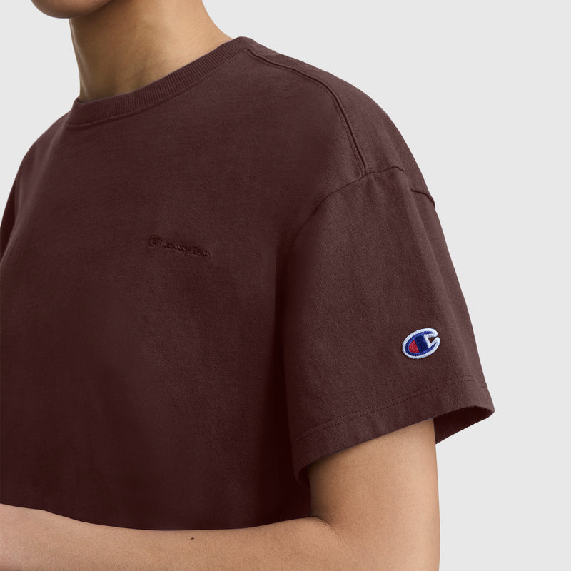 A person is shown from the shoulders down wearing the Champion Short Sleeve Loose Fit T-Shirt in maroon, with the embroidered Champion logo visible on the sleeve. The t-shirt has a relaxed fit, and the background is plain and light-colored.
