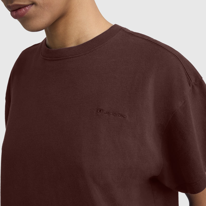 A person is shown from the shoulders up against a plain light background, wearing a maroon Champion Short Sleeve Loose Fit T-Shirt with an embroidered logo on the chest. This women’s t-shirt features a comfortable, loose fit ideal for everyday wear.