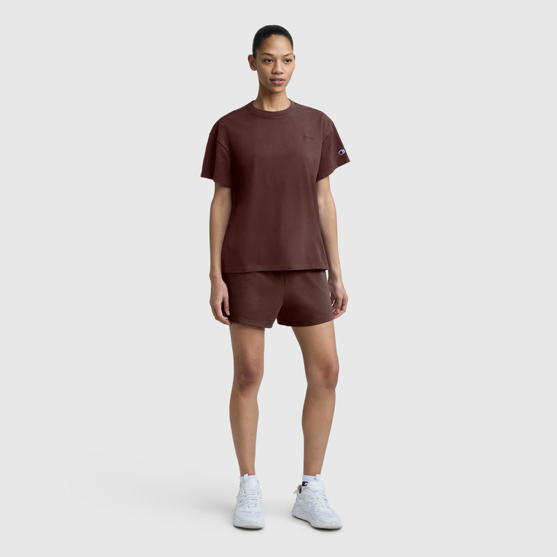 A person stands against a plain gray background, wearing the Champion Short Sleeve Loose Fit T-Shirt in brown, matching brown shorts, and white sneakers. They face forward with arms relaxed by their sides.