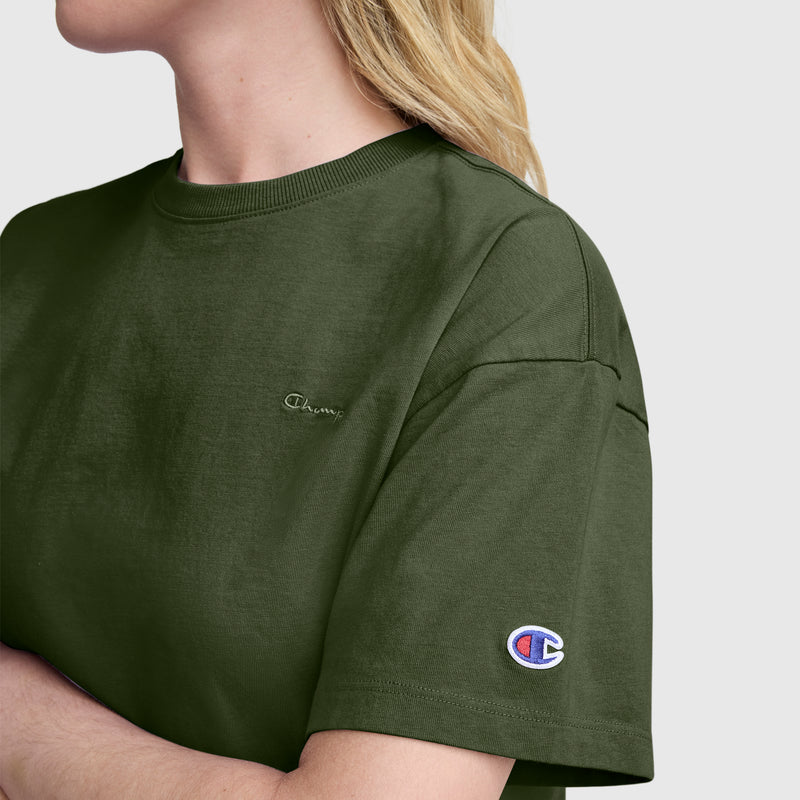 A person with blonde hair wears a Champion Short Sleeve Loose Fit T-Shirt in olive green, made from soft cotton jersey. The small Champion logo is featured on the chest, while the brand’s logo patch accents the sleeve. This relaxed tee stands out against a plain light gray background.