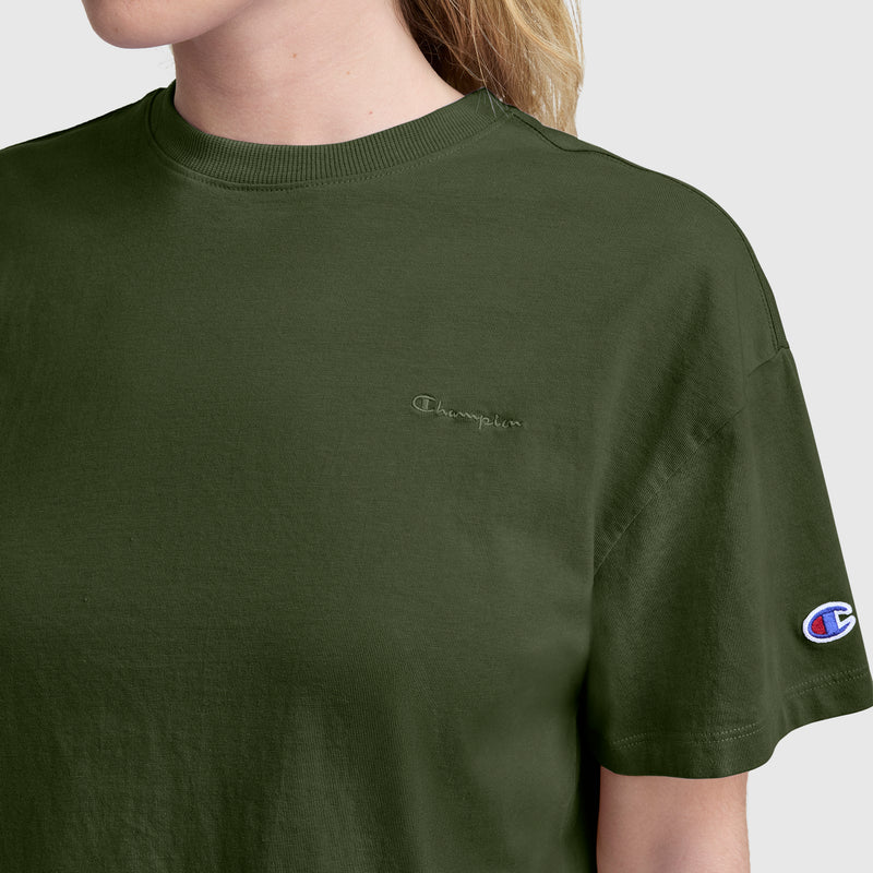 A person wearing a Champion Short Sleeve Loose Fit T-Shirt in olive green stands against a plain background, highlighting the soft cotton jersey fabric and the iconic embroidered Champion logo on the chest, along with the signature Champion patch on the left sleeve. This relaxed-fit tee delivers casual comfort and timeless style.