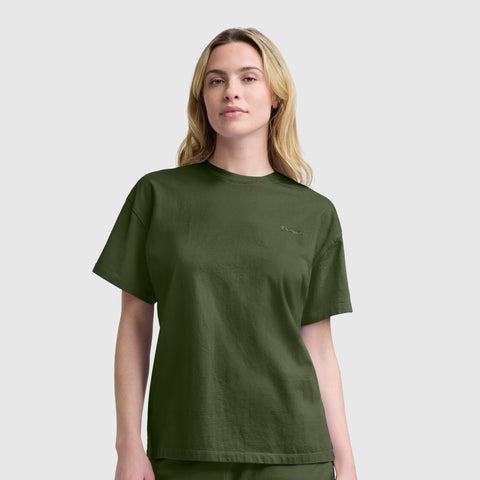 A woman with long blonde hair wears a Champion Short Sleeve Loose Fit T-Shirt in dark green, crafted from soft cotton jersey, paired with matching pants, and stands against a plain light gray background.