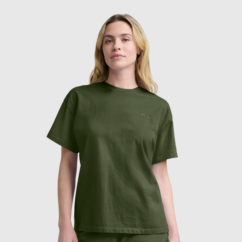 A woman with long blonde hair wears a Champion Short Sleeve Loose Fit T-Shirt in dark green, crafted from soft cotton jersey, paired with matching pants, and stands against a plain light gray background.
