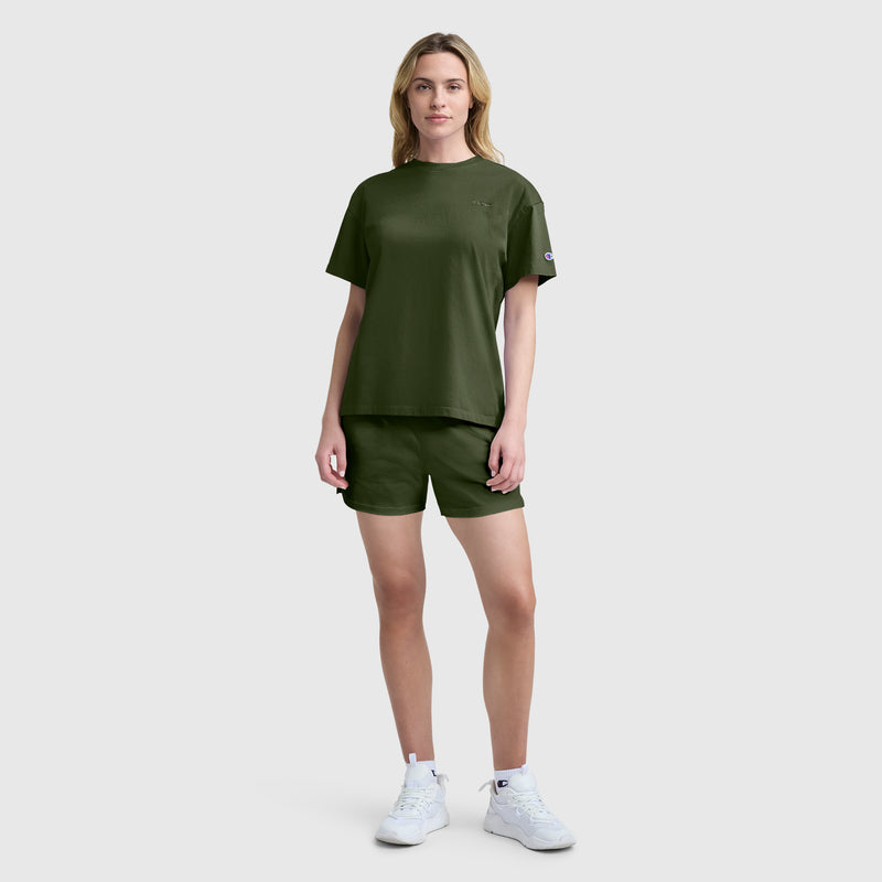 A person stands facing forward, wearing a Champion Short Sleeve Loose Fit T-Shirt in olive green paired with matching shorts and white athletic shoes, set against a plain light gray background.