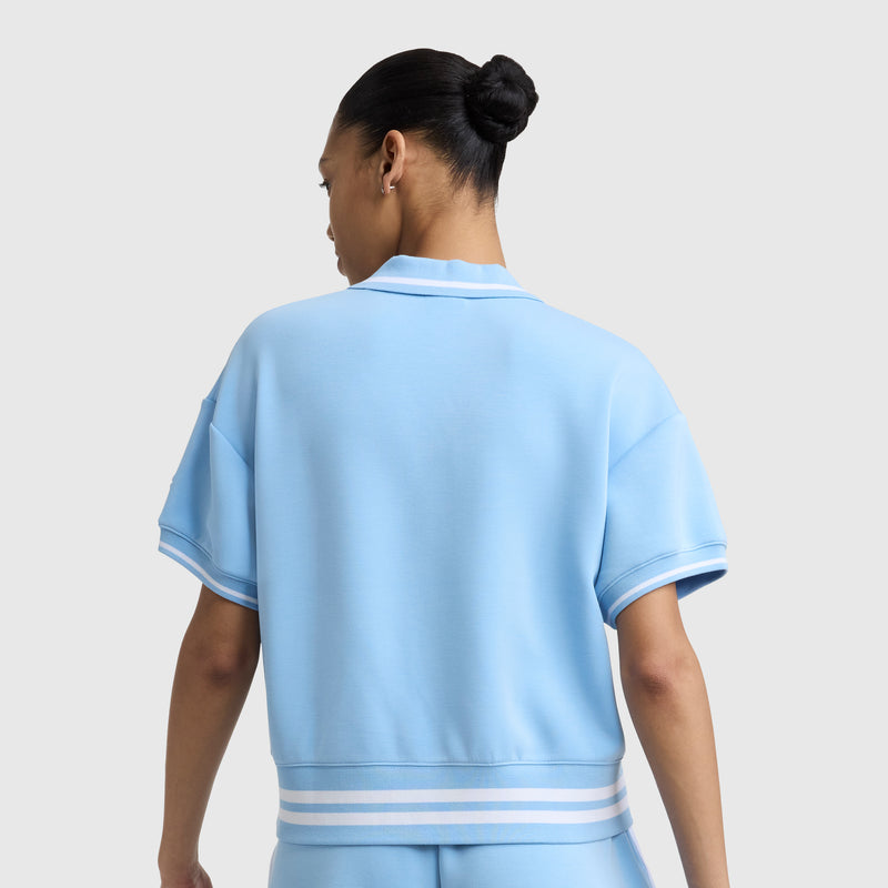 A person with dark hair in a neat bun is seen from behind wearing the Champion Tennis Court Polo, featuring an embroidered script logo and white and blue striped trim, against a plain light gray background.