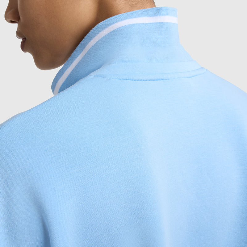 Close-up of a woman wearing the Champion Tennis Court Polo, Embroidered Script Logo, in light blue with a white collar stripe. Her face is turned away, showcasing the polo’s stylish back and neckline.