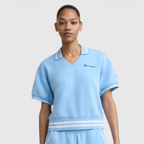 A woman wears the Champion Tennis Court Polo, featuring a light blue short-sleeve design with a white striped collar and waistband, plus an embroidered script logo, standing against a plain light gray background.