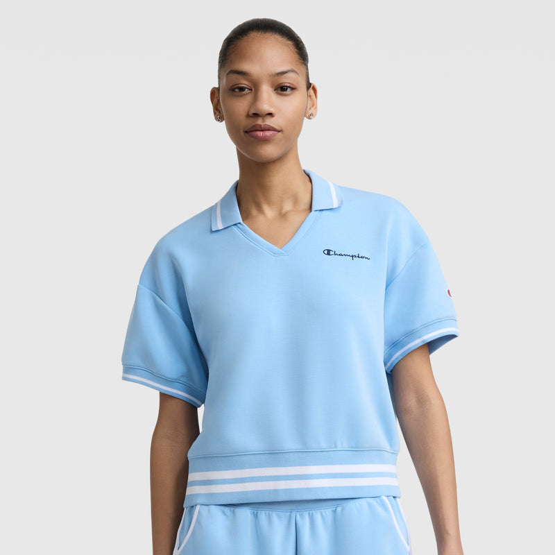 A woman wears the Champion Tennis Court Polo, featuring a light blue short-sleeve design with a white striped collar and waistband, plus an embroidered script logo, standing against a plain light gray background.