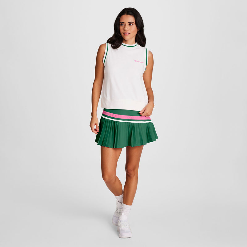 A woman stands against a plain background wearing the Champion Country Club Sleeveless Top with an embroidered script logo and a pleated green skirt with pink and white trim, white socks, and sneakers. She looks to the side, smiling slightly.
