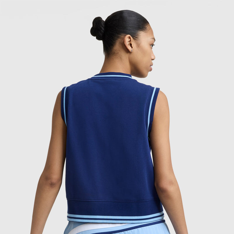A woman with dark hair in a bun is shown from behind, wearing the Champion Country Club Sleeveless Top with embroidered script logo and a light blue pleated skirt, standing out against a plain white background.