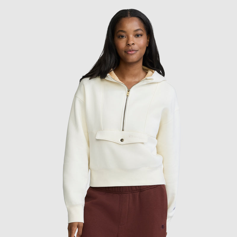 A woman stands facing forward with a neutral expression against a plain light background, wearing a cream-colored Champion Training Half-Zip Fleece Hoodie and maroon pants.