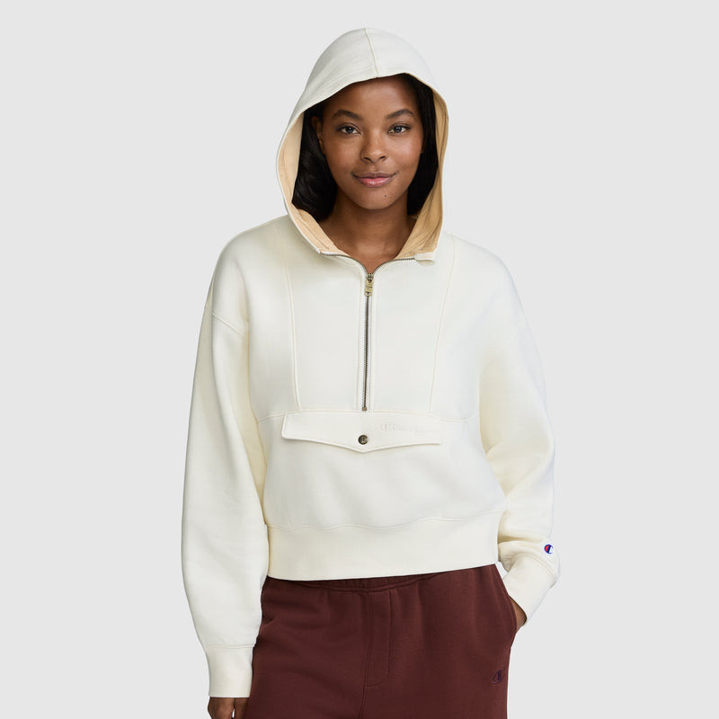 A woman stands against a plain light gray background wearing a Champion Training Half-Zip Fleece Hoodie in cream, which features a front zipper and pocket, paired with maroon pants.