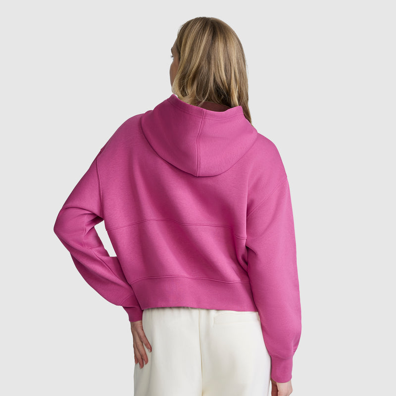 A person with long blonde hair is wearing a bright pink Champion Training Half-Zip Fleece Hoodie and white pants, standing with their back to the camera against a plain light gray background.