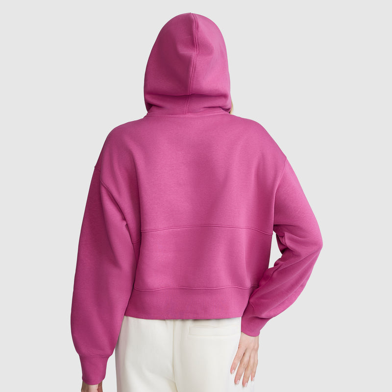 A person is seen from the back, wearing a vibrant pink Champion Training Half-Zip Fleece Hoodie with the hood up and white pants, set against a light gray background.