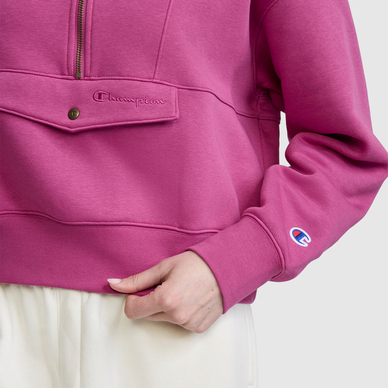 A person wearing a pink Champion Training Half-Zip Fleece Hoodie with a front pocket and white pants, hand resting near the pocket. The Champion logo is visible on the sleeve and above the pocket.