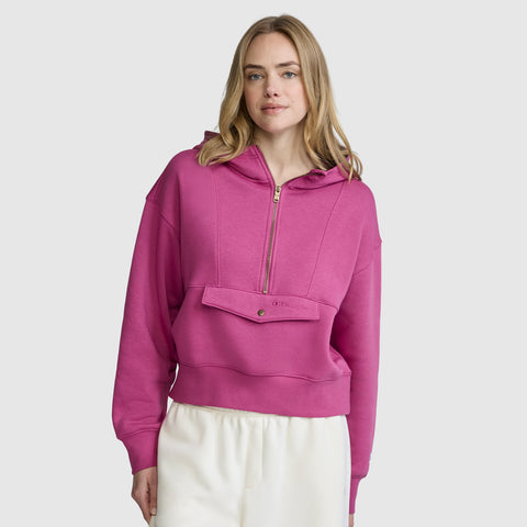 A woman wearing a bright pink Champion Training Half-Zip Fleece Hoodie and white pants stands against a plain light gray background, looking forward with a slight smile.