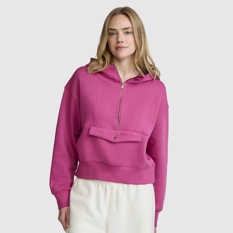 A woman wearing a bright pink Champion Training Half-Zip Fleece Hoodie and white pants stands against a plain light gray background, looking forward with a slight smile.