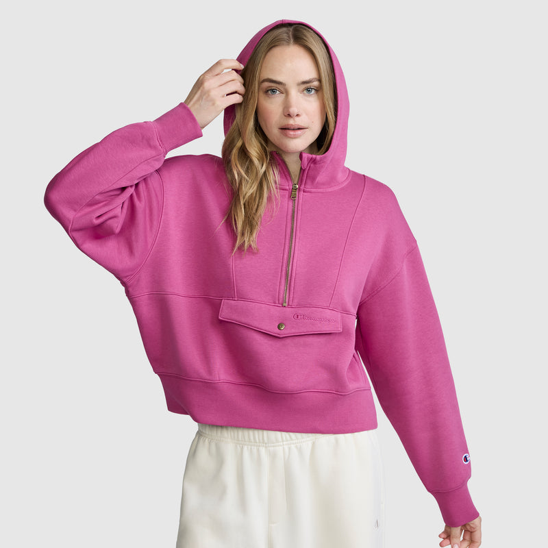 A woman poses against a plain light gray background, wearing the Champion Training Half-Zip Fleece Hoodie in bright pink with a front pocket and white pants. She holds up the hood with one hand.