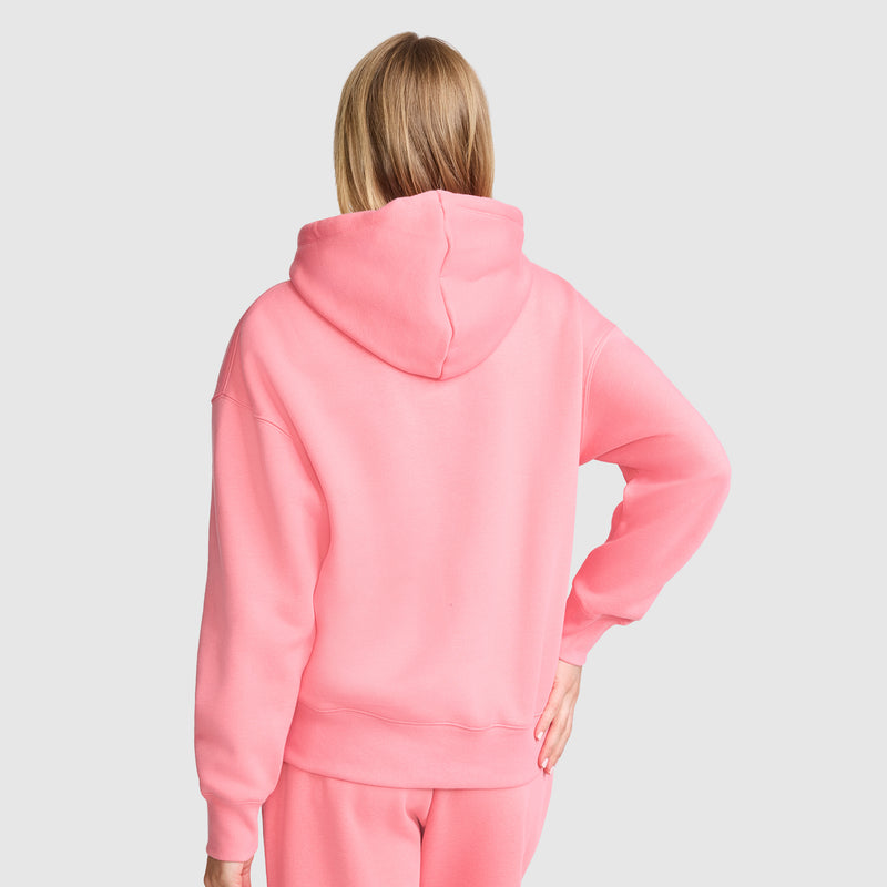 A person with blonde hair, seen from behind, wears a bright pink Champion Fleece Hoodie, Satin Applique with matching pants. Their right hand rests on their hip against a plain light gray background. The hoodie features satin applique details characteristic of the Champion brand.