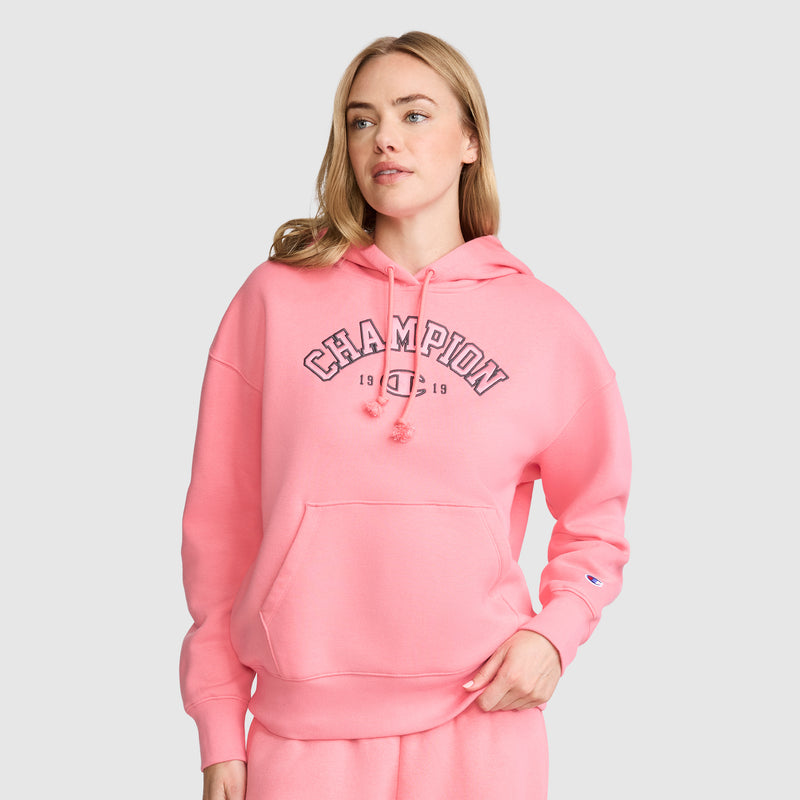 A woman stands against a plain background with one hand in her pocket, wearing the Champion Fleece Hoodie, Satin Applique and matching pants, effortlessly showcasing a modern take on varsity style while looking slightly to the side.