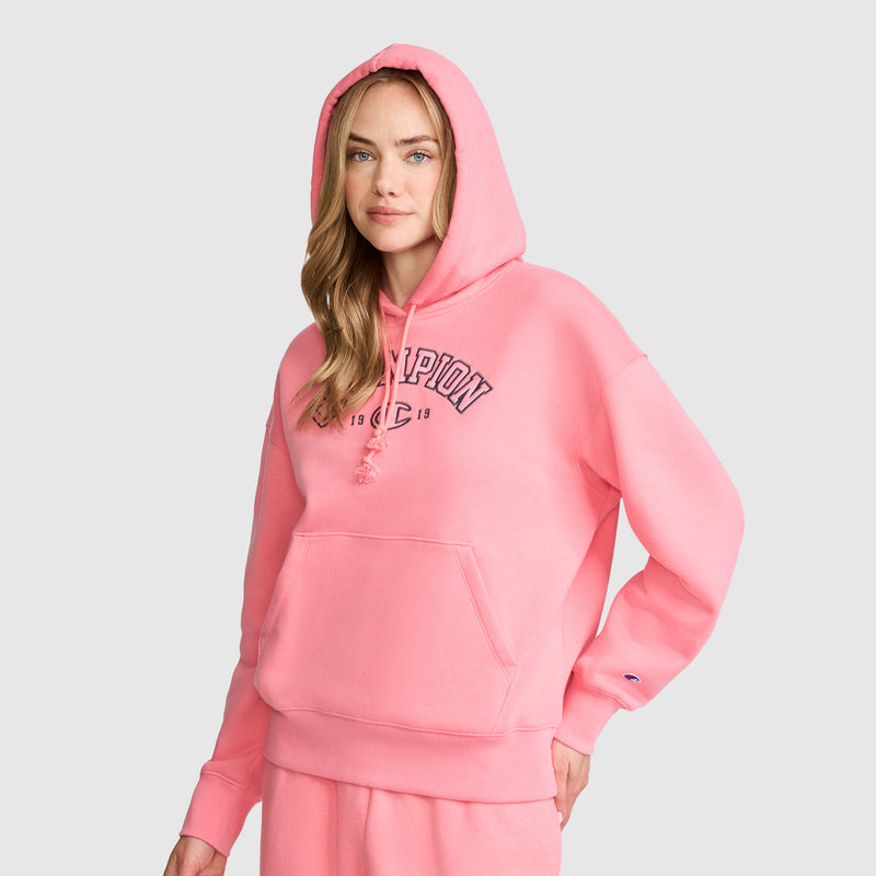 A woman with long, wavy hair stands facing forward against a plain light background, wearing the Champion Fleece Hoodie, Satin Applique in pink. The varsity-style hoodie features matching sweatpants and bold satin applique detailing for a stylish finish, with one hand tucked into her pocket.
