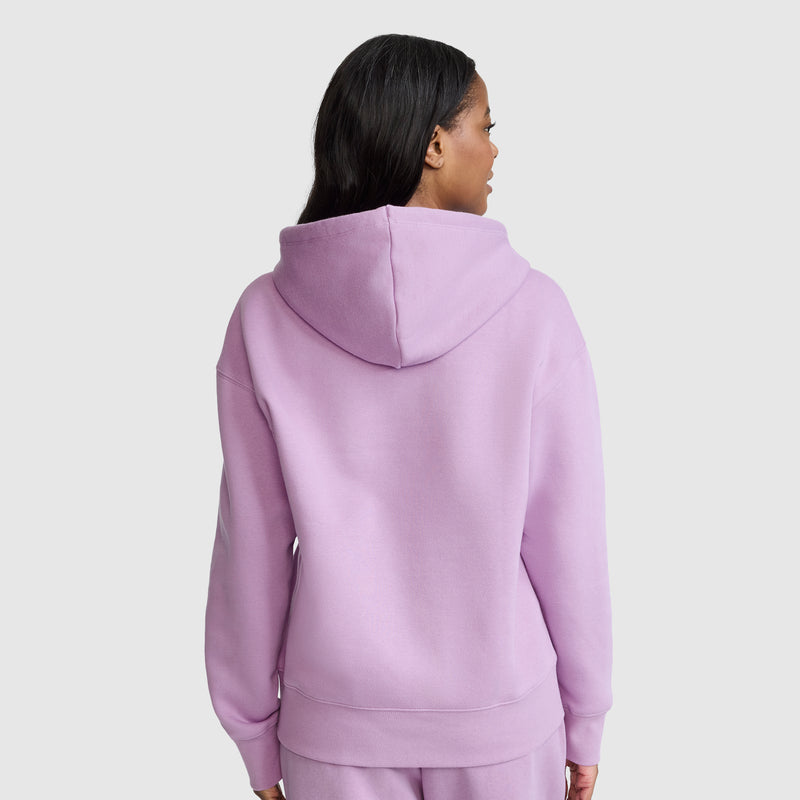 A woman with long dark hair, dressed in the Champion Fleece Hoodie with Satin Applique and matching pants in light purple, stands facing away from the camera against a plain light gray background.