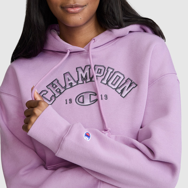 A woman wearing a light purple Champion Fleece Hoodie, Satin Applique, featuring "CHAMPION 1919" in a classic varsity style on the front and a small Champion logo on the sleeve.
