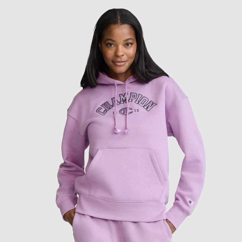 A woman stands against a plain light gray background, smiling with one hand in the pocket of her light purple Champion Fleece Hoodie, Satin Applique varsity style hoodie and matching pants.