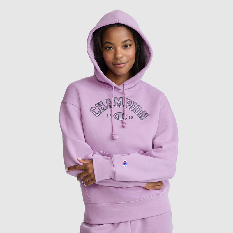 A woman wearing a lavender Champion Fleece Hoodie, Satin Applique, with the hood up stands against a light gray background, arms crossed and smiling slightly.
