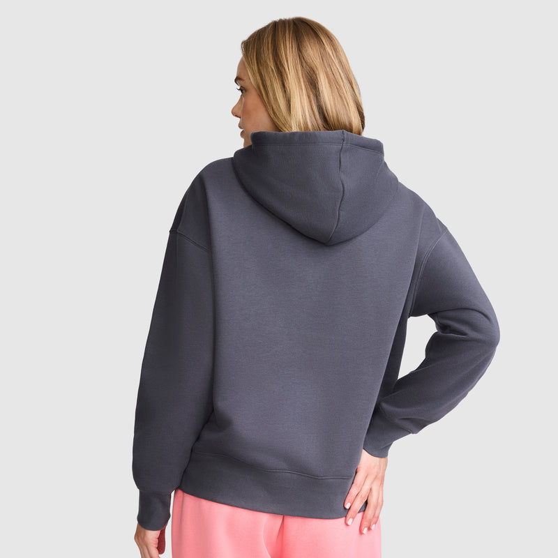 A woman with blonde hair wears a Champion Fleece Hoodie, Satin Applique in dark gray and pink pants, standing with her back to the camera and one hand on her hip against a light gray background.