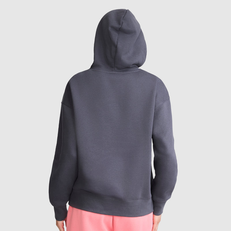 A person is seen from the back against a plain light gray background, wearing a Champion Fleece Hoodie with satin applique in dark gray, hood up, paired with light pink pants.