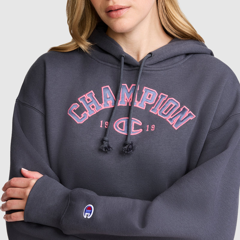 A woman with light brown hair crosses her arms while wearing a Champion Fleece Hoodie, Satin Applique in dark gray. “CHAMPION 1919” appears across the front in pink and white letters, and the signature Champion logo patch is featured on the sleeve, giving this hoodie its classic varsity style.