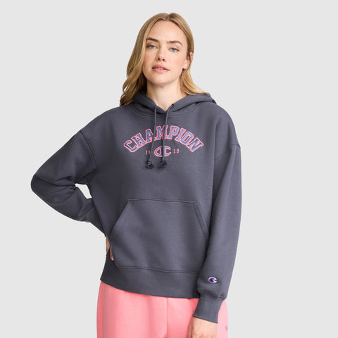 A woman with long blonde hair wears a dark gray Champion Fleece Hoodie, Satin Applique, featuring pink varsity-style lettering and light pink sweatpants, standing against a plain light gray background.