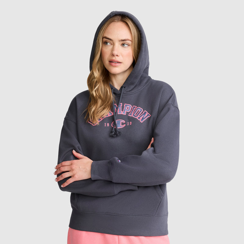 A woman with long blonde hair wears a dark gray Champion Fleece Hoodie, Satin Applique, featuring varsity-style pink lettering. She pairs it with light pink pants and stands with her arms crossed against a plain light background.