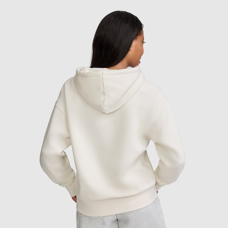 A person with long dark hair is wearing a Champion Fleece Hoodie, Satin Applique in an off-white shade and light gray pants, standing with their back to the camera against a plain light background.