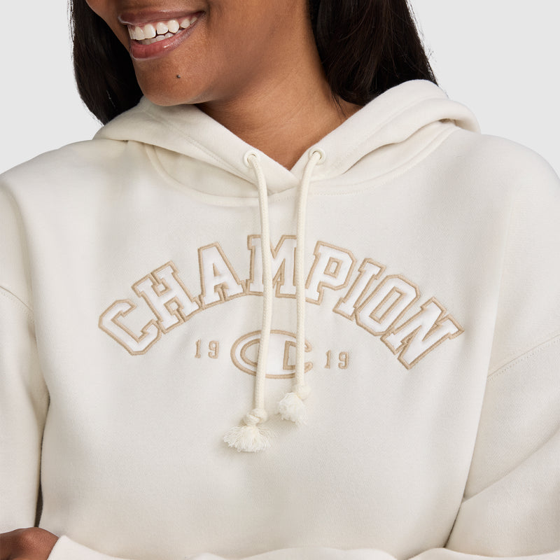 A smiling person wearing an off-white Champion Fleece Hoodie, Satin Applique, featuring CHAMPION 1919 embroidered in beige letters on the chest for a subtle varsity-inspired look.