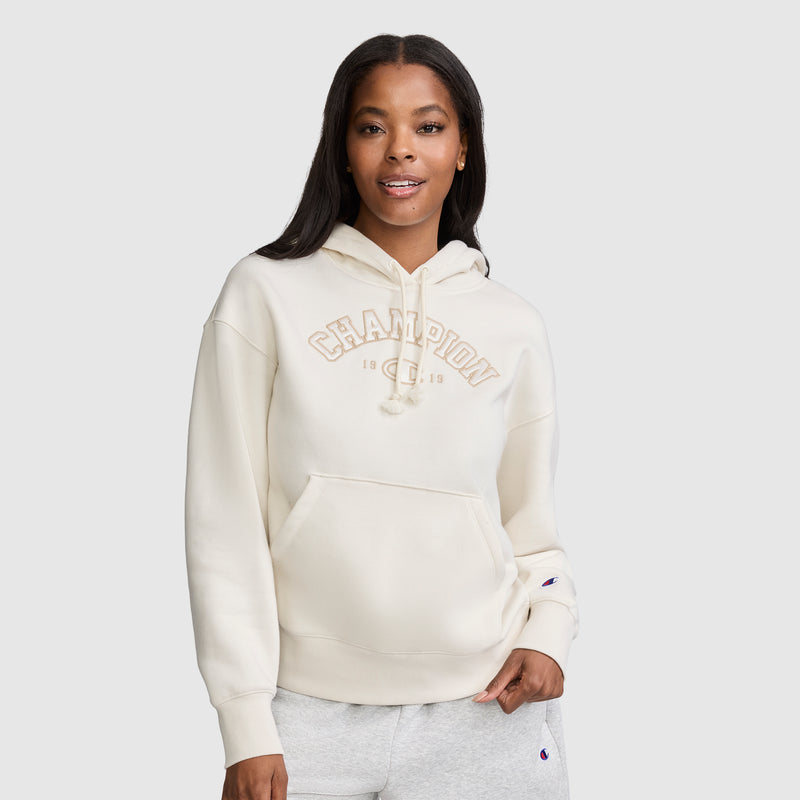 A woman stands against a plain background, smiling with one hand in her pocket, wearing a cream-colored Champion Fleece Hoodie with satin applique "CHAMPION" lettering on the front.