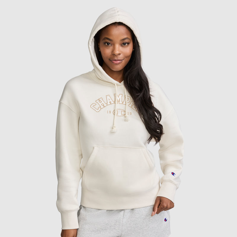 A woman with long dark hair and a relaxed expression stands against a plain light gray background, wearing the Champion Fleece Hoodie, Satin Applique in cream with the hood up. One hand is in her pocket as she showcases the varsity style hoodie featuring classic satin applique detailing for an elevated casual look.