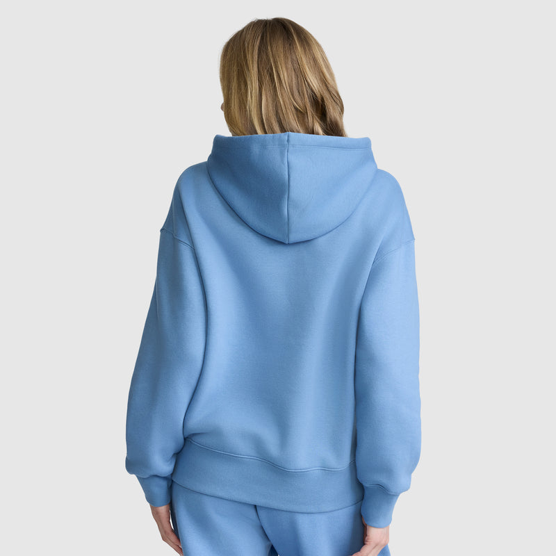 A person with blonde hair is seen from behind, wearing the Champion Fleece Hoodie with satin applique and matching sweatpants, standing against a plain white background.