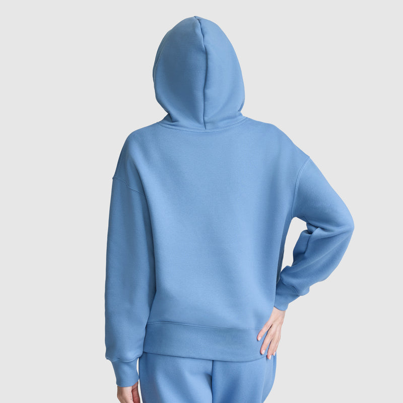 A person wearing a blue Champion Fleece Hoodie with satin applique and matching pants stands facing away, one hand on their hip. The hood is up, covering their head, against a plain light background.