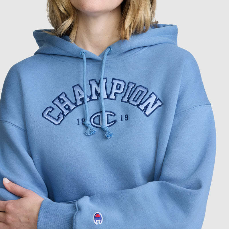 A woman wearing a light blue Champion Fleece Hoodie, Satin Applique, featuring classic varsity style and CHAMPION 1919 text on the front, stands with her arms crossed against a plain background.