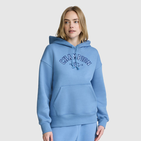 A woman wearing a light blue Champion Fleece Hoodie, Satin Applique with a front pocket and matching sweatpants stands against a plain light background. This varsity-inspired hoodie from Champion blends comfort and classic athletic style for an effortlessly cool look.