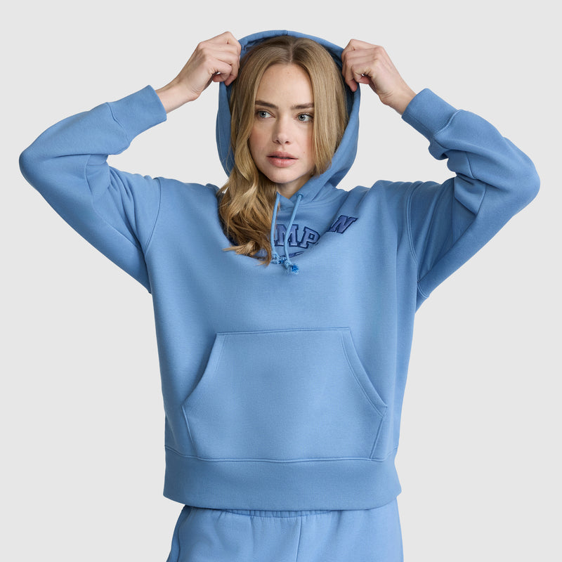 A woman wears the Champion Fleece Hoodie with Satin Applique and matching sweatpants, pulling up her hood as she faces forward against a plain background, with one hand on each side of the hood.