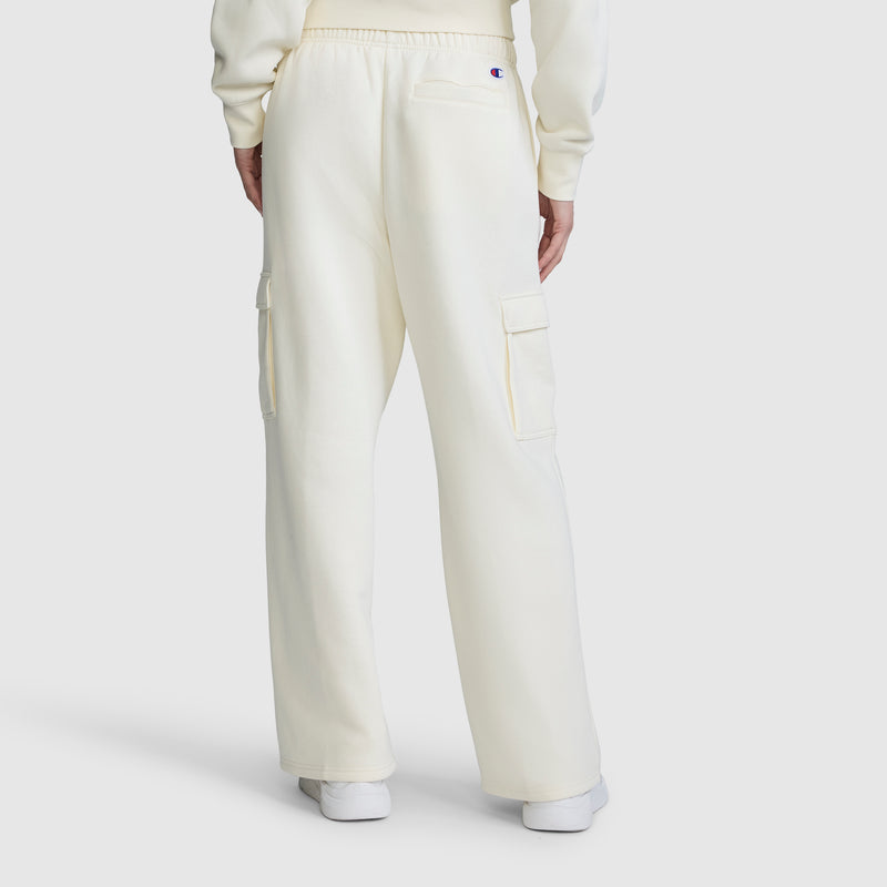A person is seen from the back, standing against a plain white background, wearing the Champion Fleece Cargo Training Pant in a cream color paired with a matching sweatshirt. The pants feature side and back pockets, delivering comfort and utility for everyday wear.
