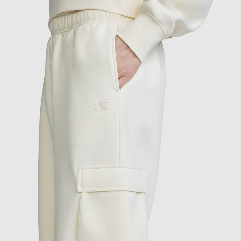 A person wearing the Champion Fleece Cargo Training Pant in off-white with a hand in the pocket. These pants showcase an embroidered C logo and a utility pocket with a flap on the thigh, combining style and functionality.