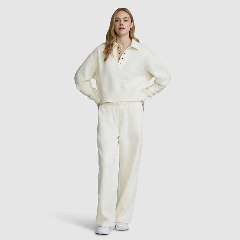 A person with long hair faces forward, wearing white shoes and a cream-colored pullover paired with Champion Fleece Cargo Training Pant. The background is plain white.