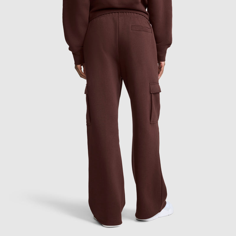 A person is shown from the back, standing against a plain light background, wearing Champion Fleece Cargo Training Pants in brown with a matching top and white shoes. The pants feature side and back pockets and offer a relaxed fit.
