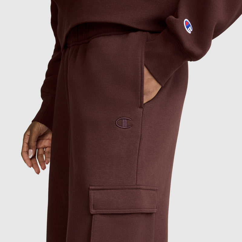 A person wearing Champion Fleece Cargo Training Pant sweatpants in maroon and a matching sweatshirt, with one hand in their pocket. The Champion logo is displayed on both the pants and the sweatshirt sleeve.